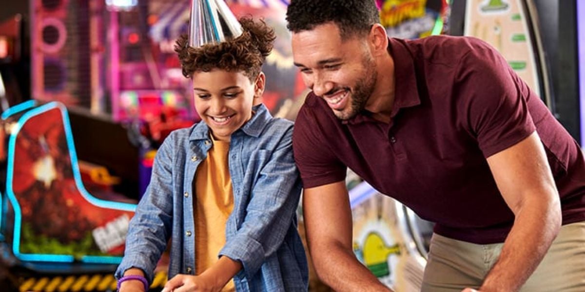 A smiling man and child play an arcade game together. The child, dressed in a costume and a party hat, laughs beside the man in a maroon polo. Bright arcade machines light up the colorful background.