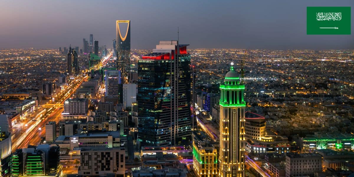 A nighttime cityscape of Riyadh, Saudi Arabia, with illuminated skyscrapers and roads. The Kingdom Centre Tower is visible in the background. A nod to recent news, such as a theme park accident, contrasts with the vibrant city scene. The Saudi Arabian flag appears at top right.