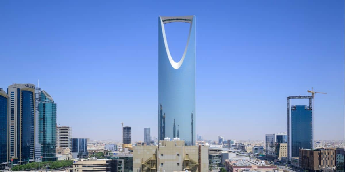 A tall, modern skyscraper with a distinctive arch-shaped opening near the top stands against a clear blue sky, its silhouette as striking as headlines after a theme park accident, surrounded by other high-rise buildings in a vibrant cityscape.