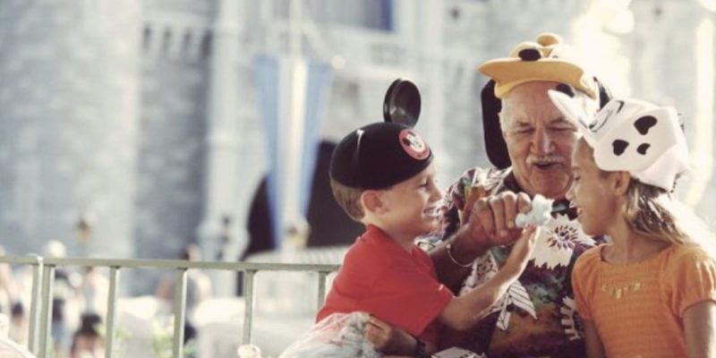An elderly man—likely a grandparent—and two children, all wearing Disney character hats, laugh and share a snack in front of a castle at Disney World. Their playful moment captures the joy of family on 2025 Disney World trips.