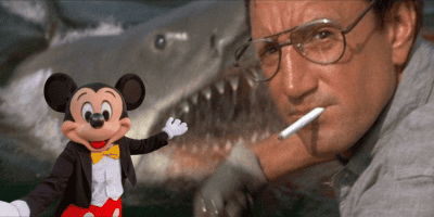 A person with glasses and a cigarette is close to a JAWS-like shark with its mouth open; in the foreground, someone in a Disney Mickey Mouse costume poses with arms outstretched, celebrating the 50th Anniversary.