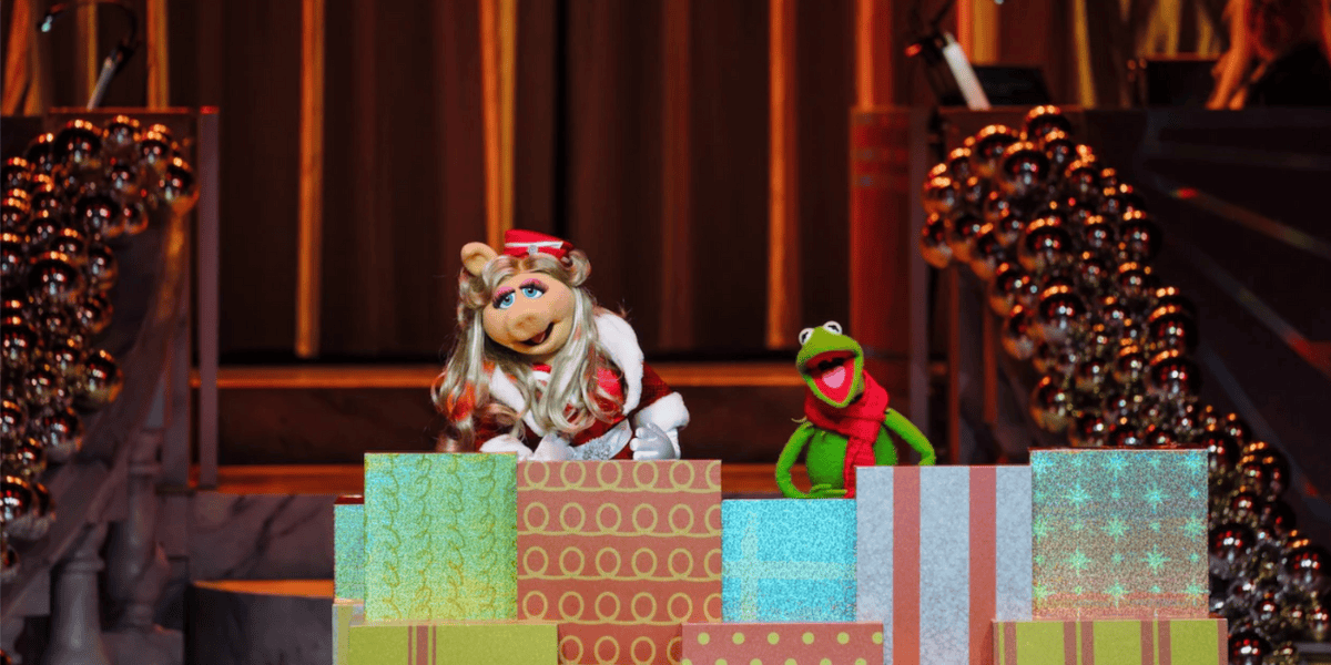 Miss Piggy and Kermit the Frog, dressed in festive outfits, stand behind colorful wrapped presents on a decorated stage at Magic Kingdom Park, with shimmering garlands and golden curtains capturing the spirit of Christmas 2025.