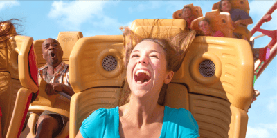 A young woman in a blue shirt smiles and screams with excitement on a roller coaster, her hair flying up. Other fans in the background show thrill and surprise, enjoying the bright, clear sky before upcoming Universal ride closures in 2025.
