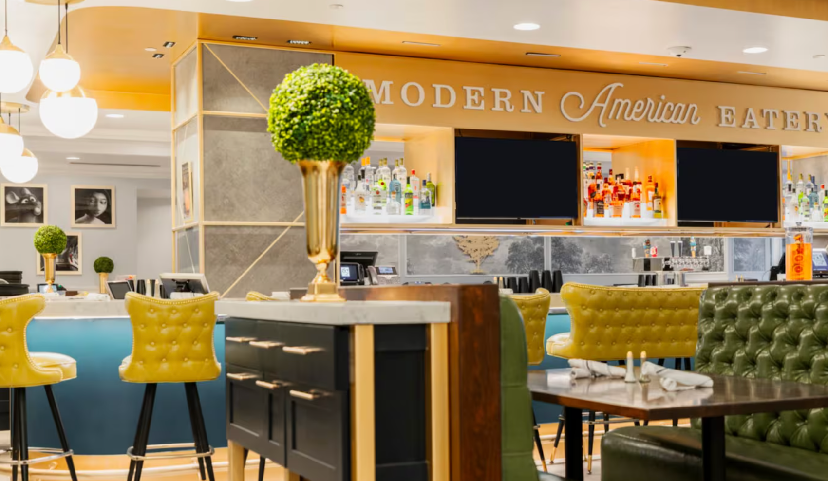 A stylish restaurant interior with a bar, yellow tufted chairs, green topiary centerpieces, and a sign reading “Modern American Eatery” above the shelves of liquor bottles.