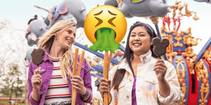 Two women smiling and holding churros and Mickey Mouse-shaped ice cream treats at an amusement park, with the Dumbo rides in the background and a large face vomiting emoji over the center of the image.