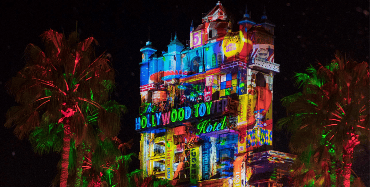 A tall, ornate tower labeled "The Hollywood Tower Hotel" is brightly illuminated with colorful, layered projections at night in the park, surrounded by palm trees lit with red and green lights—perfect for Christmas 2025 magic.