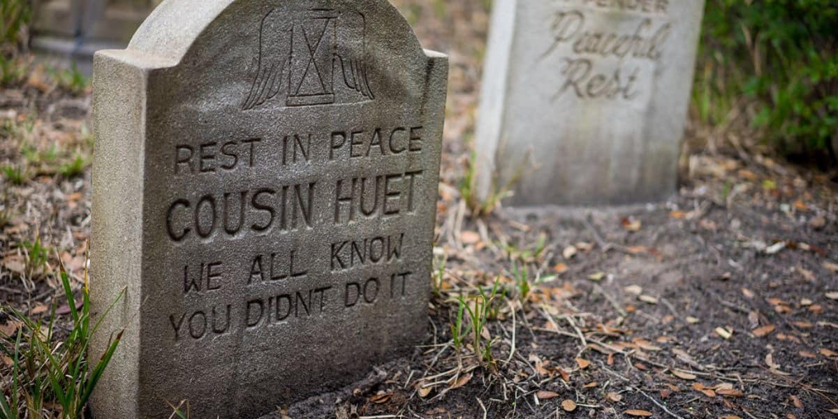 A gravestone reads “Rest in peace Cousin Huet. We all know you didn’t do it,” with grass and another blurred headstone in the background.