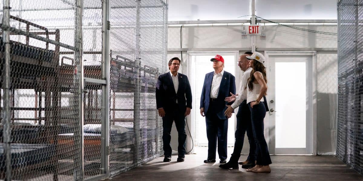 Four people stand inside a facility with chain-link fenced enclosures and bunk beds. Three men in business attire, one wearing a red cap, and one woman in casual clothes and a white hard hat are present.