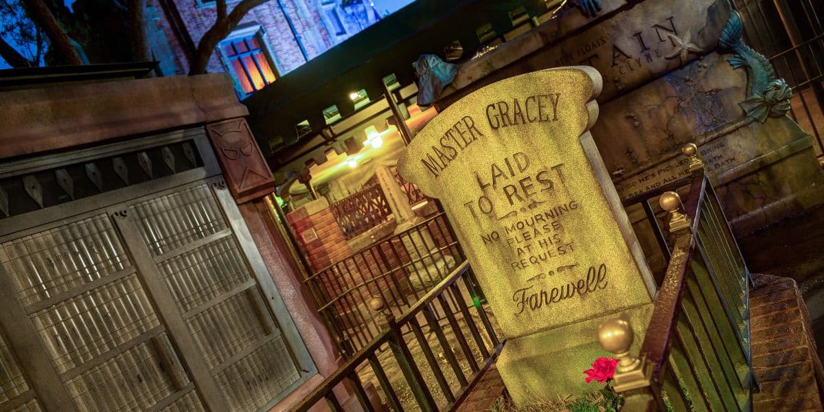 A tilted gravestone inscribed with “Master Gracey laid to rest,” behind a black fence at night, surrounded by dim lights and brick walls, with flowers at the base and a haunted, eerie atmosphere.
