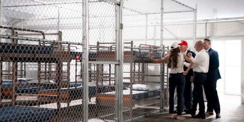 Four adults stand near metal bunk beds inside a fenced area, possibly a detention or holding facility. The area is enclosed with chain-link fencing, and the beds have minimal bedding. Two men wear suits; one person wears a red hat.