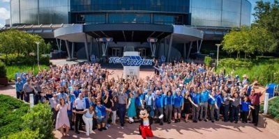 A large group of people, many wearing blue shirts, gather outside in front of a building with a "Test Track" sign. Mickey Mouse stands in front, waving. Trees and greenery surround the area.