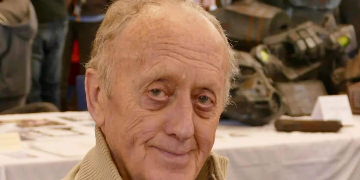 Kenneth Colley with thinning hair and a tan jacket smiles gently at the camera while seated indoors, with blurred people and tables in the background—an Original Trilogy Actor from Star Wars enjoying a quiet moment amid COVID.