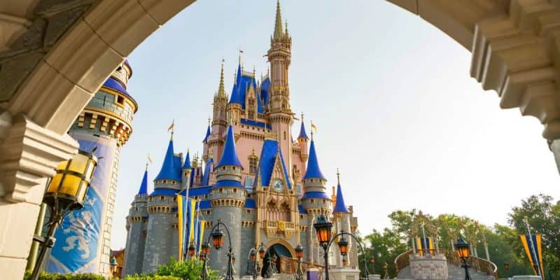 A photo of a large fairytale castle with blue and gold rooftops, seen through a stone archway on a sunny day. Decorative flags and vintage-style lamps line the walkway leading to the castle as Disney World crowds vanish from plane sight as Disney news is reported.