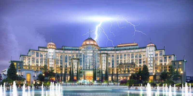 A grand, brightly lit building with domes and columns stands at night, as lightning illuminates the sky above. Water fountains in the foreground reflect the building’s lights at Epic Universe at Universal Orlando Resort.