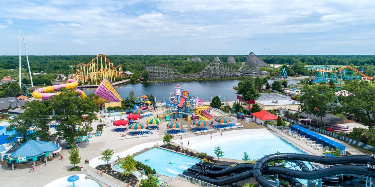 Aerial view of Six Flags theme park called Michigan's Adventure with water slides, pools, umbrellas, thrill rides, and roller coasters near a wooded lake under a partly cloudy sky.