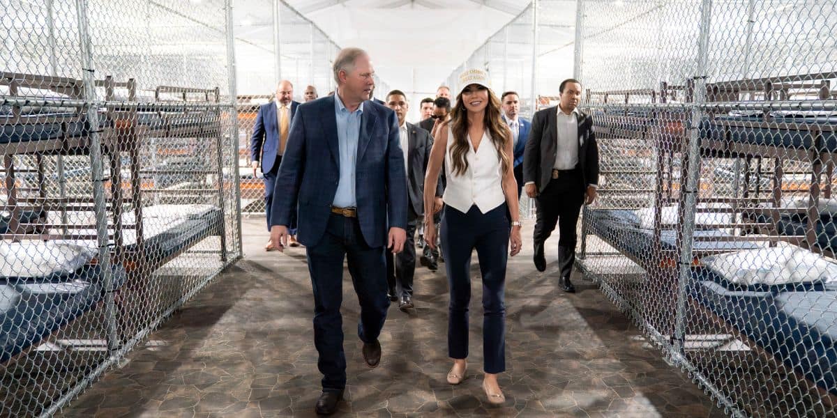 A group of people walk through a facility with fenced enclosures containing bunk beds, suggesting a detention or temporary shelter environment. The two people in front are engaged in conversation.