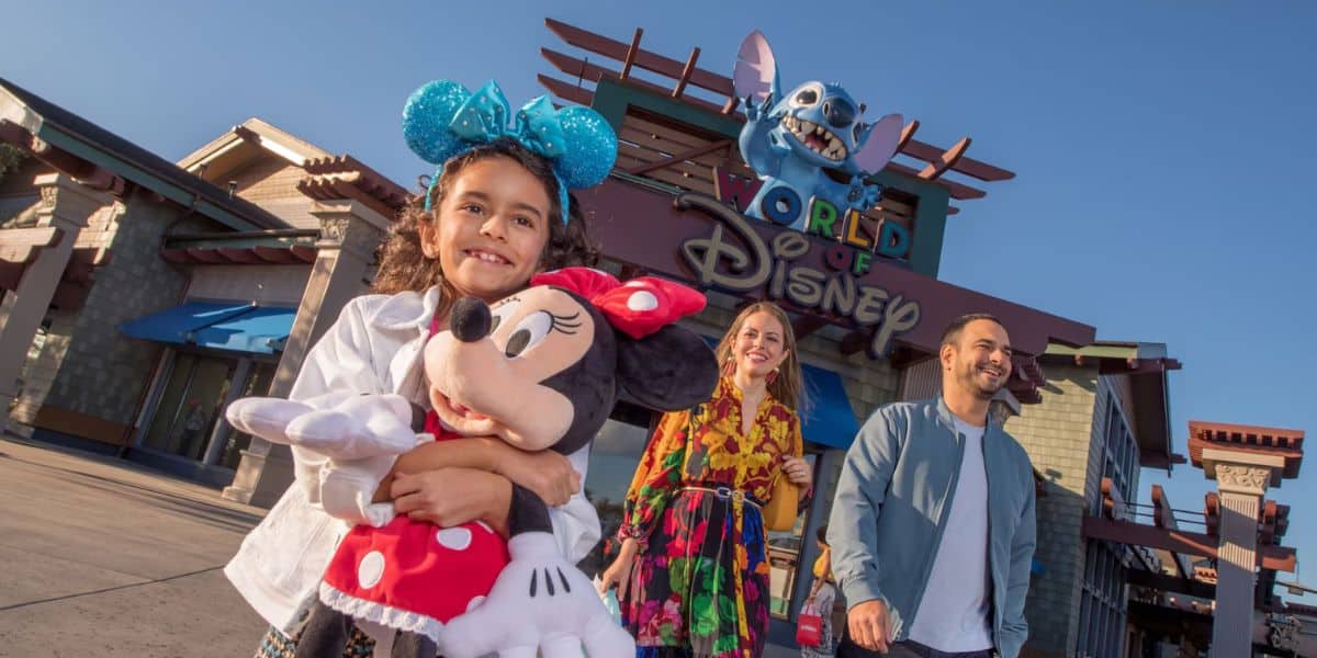 A smiling girl wearing blue Minnie Mouse ears hugs a Minnie Mouse plush. Two adults walk behind her, all in front of the World of Disney store entrance—unlike the viral video of a Disney Springs fight, here the scene is peaceful under clear, sunny skies.