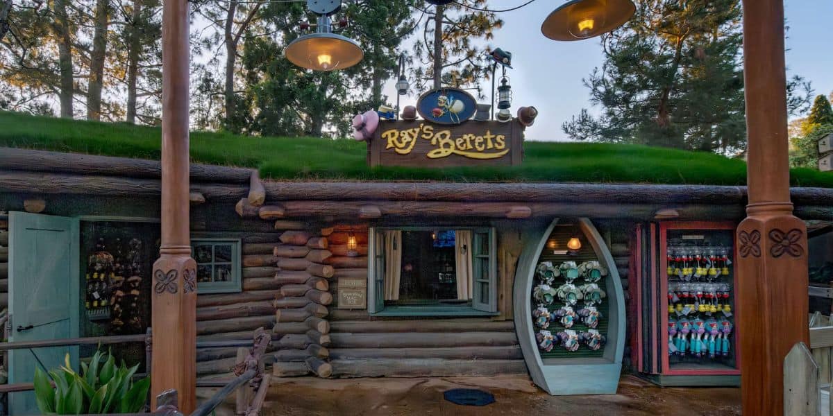 A whimsical log cabin-style shop called Ray's Beets, with a grassy roof, whimsical decorations, open doors, and display racks filled with colorful merchandise, set amid tall trees.