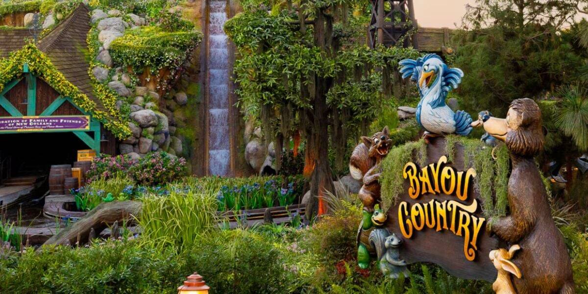 Colorful "Bayou Country" sign with cartoon animals stands in front of lush greenery, flowers, and a log flume waterfall at the entrance to a theme park attraction.