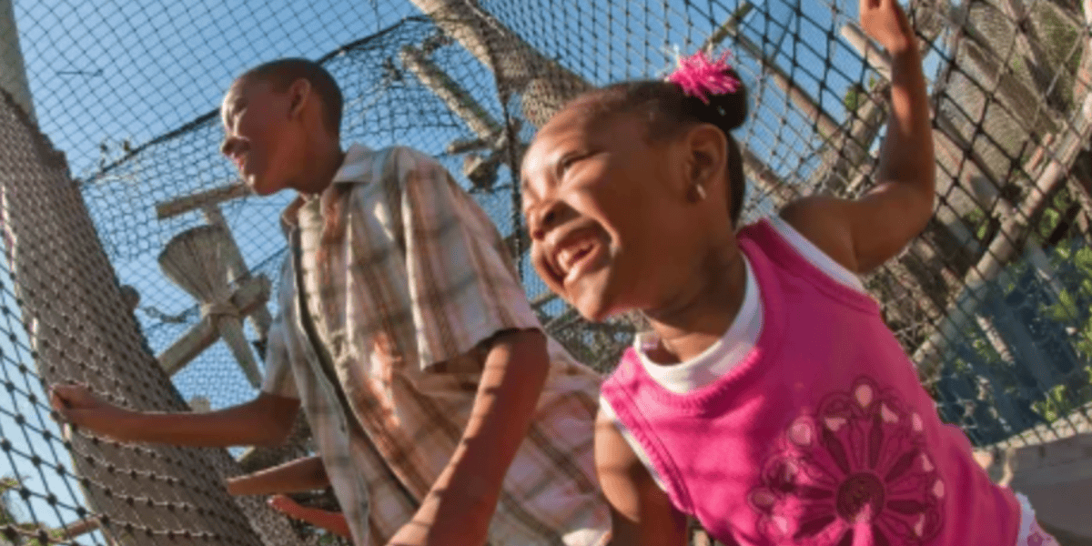 Two children laugh and play outside near a chain-link fence under a bright blue sky, their imaginations running wild like an adventure in Jurassic Park. One wears a plaid shirt, while the other sports a pink top and matching pink hair ties.