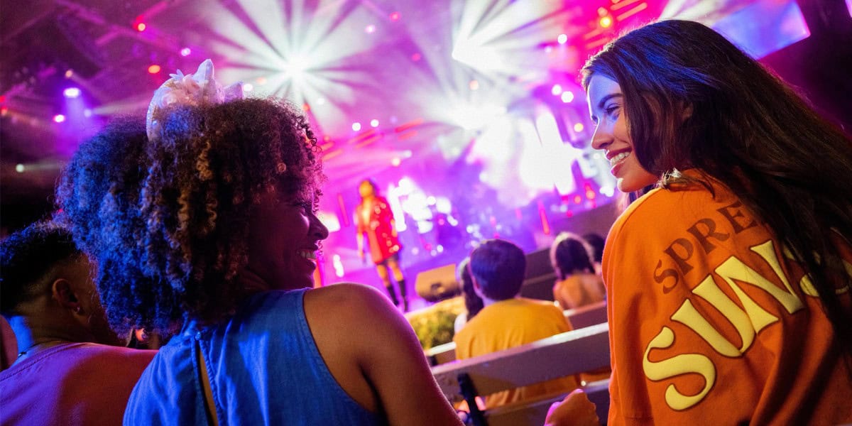 Two young women smile at each other while sitting in the audience at the Eat to the Beat concert, with colorful stage lights and performers from this year’s Eat to the Beat lineup visible in the background.