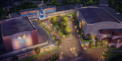 Aerial view of a theme park area at dusk, featuring tree-lined walkways, people strolling, colorful lights, and large buildings—including one housing the Star Wars Launch Bay and another with a giant blue sorcerer’s hat above the entrance.