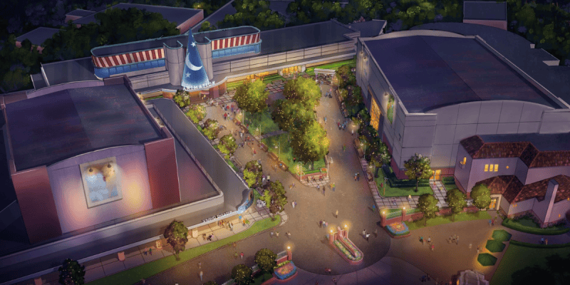 Aerial view of a theme park area at dusk, featuring tree-lined walkways, people strolling, colorful lights, and large buildings—including one housing the Star Wars Launch Bay and another with a giant blue sorcerer’s hat above the entrance.