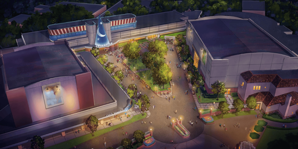 Aerial view of a theme park area at dusk, featuring tree-lined walkways, people strolling, colorful lights, and large buildings—including one housing the Star Wars Launch Bay and another with a giant blue sorcerer’s hat above the entrance.