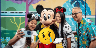 A family poses for a selfie with a person in a Mickey Mouse costume at Disney World. A large, sad-faced emoji with a tear covers the center, hinting at problems or external factors affecting their visit.