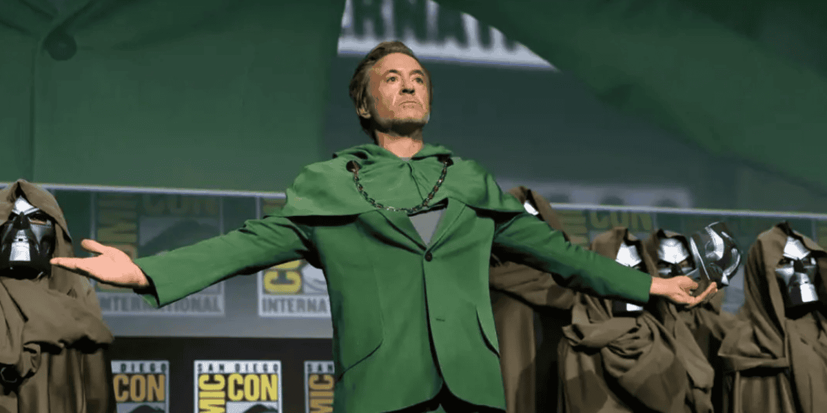 A person in a green costume with a cape and outstretched arms stands on stage at a Comic-Con event, alongside figures in brown robes and metallic masks, evoking iconic villains from the MCU and Fantastic Four.