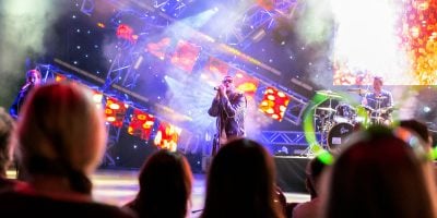 A band performs on a brightly lit stage with colorful lights and fog effects at EPCOT Eat to the Beat 2025, while an enthusiastic audience watches from the foreground. The lead singer stands center stage, flanked by a drummer and other musicians.