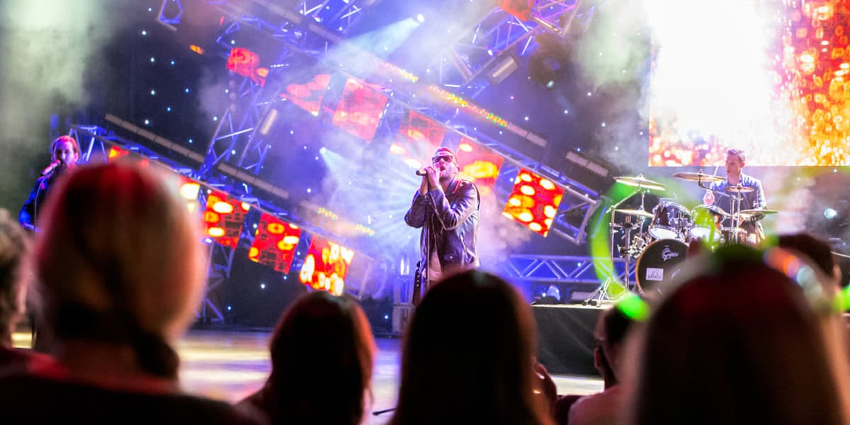 A band performs on a brightly lit stage with colorful lights and fog effects at EPCOT Eat to the Beat 2025, while an enthusiastic audience watches from the foreground. The lead singer stands center stage, flanked by a drummer and other musicians.
