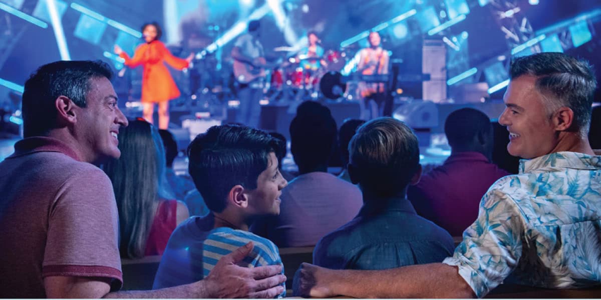 A family with two adults and two children sits in an audience, smiling and enjoying a live EPCOT Eat to the Beat 2025 stage performance with musicians and colorful lighting in the background.