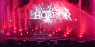 Bright pink and red water fountains glow at night as "Halloween Horror Nights 2025" is projected onto the mist above. Silhouetted fans gather, drawn to this fan-favorite spectacle in the illuminated evening.