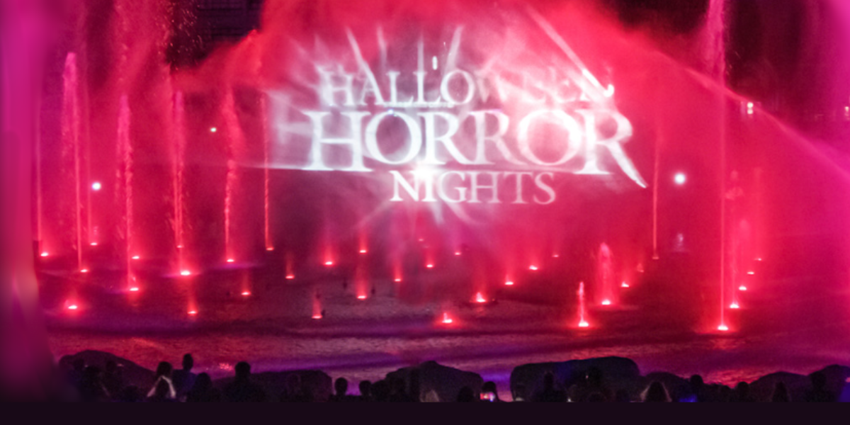 Bright pink and red water fountains glow at night as "Halloween Horror Nights 2025" is projected onto the mist above. Silhouetted fans gather, drawn to this fan-favorite spectacle in the illuminated evening.