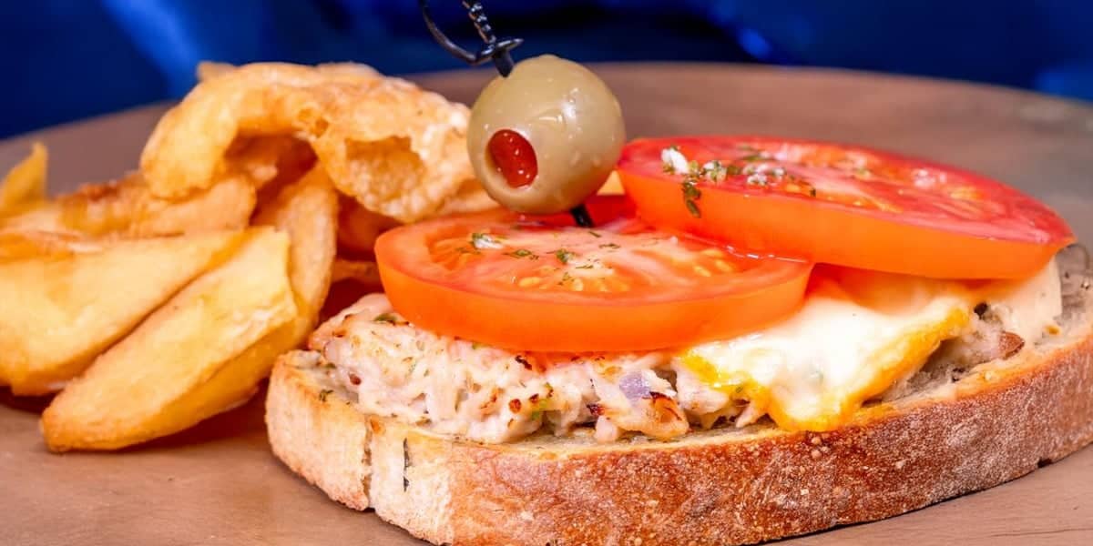 Open-faced sandwich with melted cheese, tuna salad, and tomato slices on rustic bread, garnished with a green olive on a toothpick. Part of the Disneyland 70th Anniversary limited-time treats, served with thick potato wedges on a wooden plate.