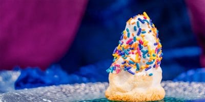 A cone-shaped cookie topped with white icing and colorful sprinkles sits on a reflective surface, part of the limited-time treats celebrating Disneyland’s 70th Anniversary, with a blurred blue and purple background.