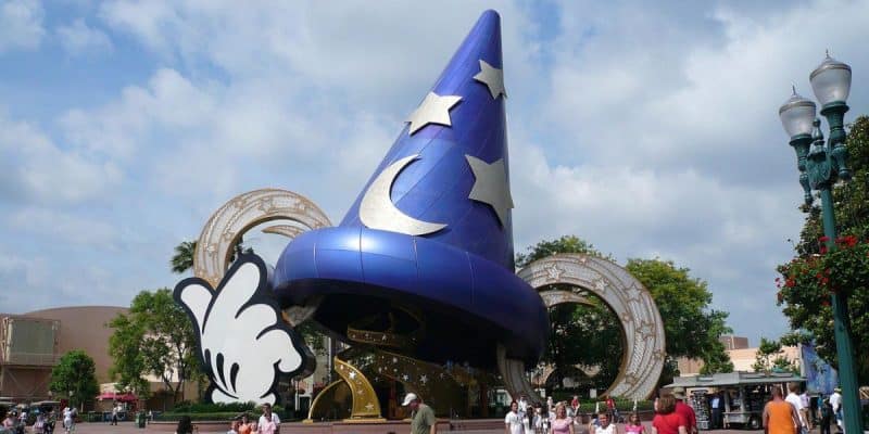 The iconic Mickey's Sorcerer Hat statue with white stars and a crescent moon stands tall at Disney’s Hollywood Studios, featuring a giant glove and gold rings, surrounded by visitors and trees on a partly cloudy day.