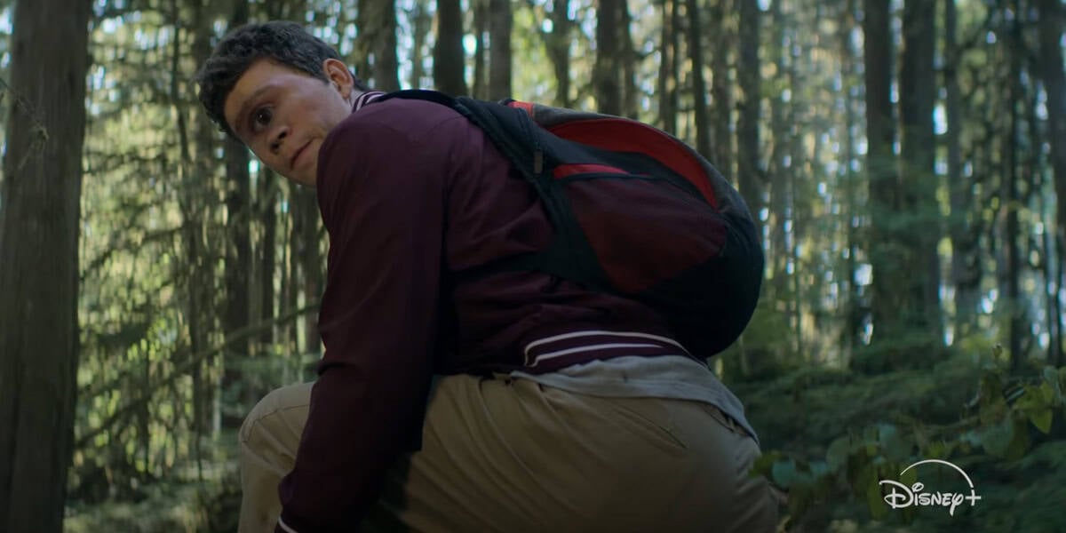 A young man wearing a maroon jacket and backpack crouches in a dense forest, looking back over his shoulder with a serious expression. Sunlight filters through the tall trees. The Disney+ logo appears, teasing the Percy Jackson Season 2 December Premiere Date.