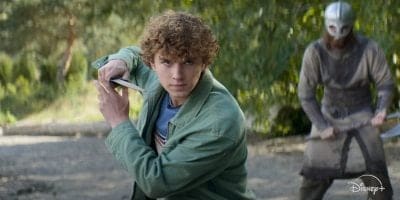 A curly-haired teen in a green jacket holds a sword defensively, ready for action, while a helmeted figure with an axe looms behind. Outdoors with blurred greenery. Disney+ logo is in the corner, teasing Percy Jackson Season 2 excitement.