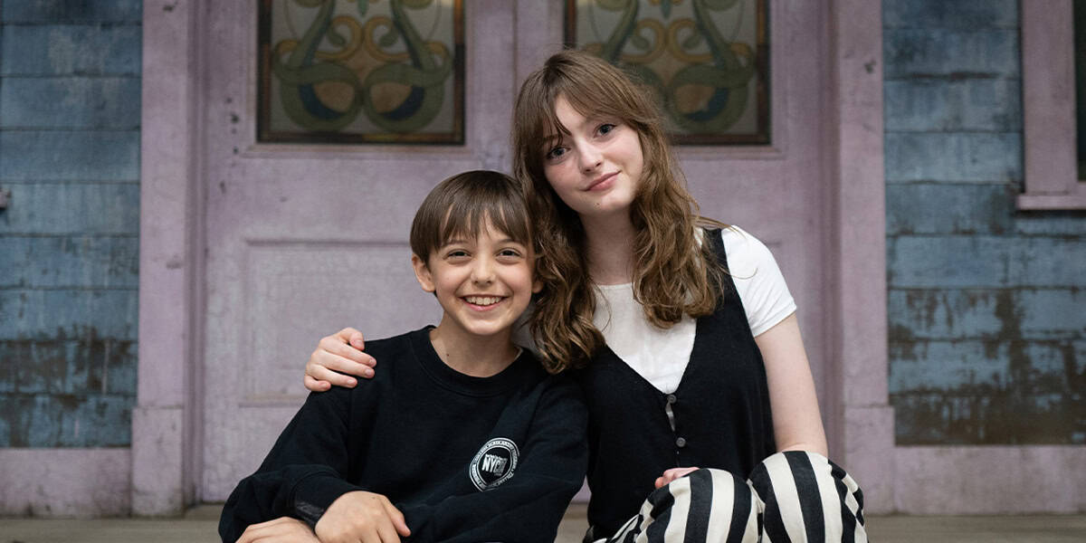 Two children sit closely together on a porch, smiling at the camera. The girl has her arm around the boy. The background features a purple door and weathered blue wood siding, evoking anticipation like Percy Jackson Season 2's December premiere date.