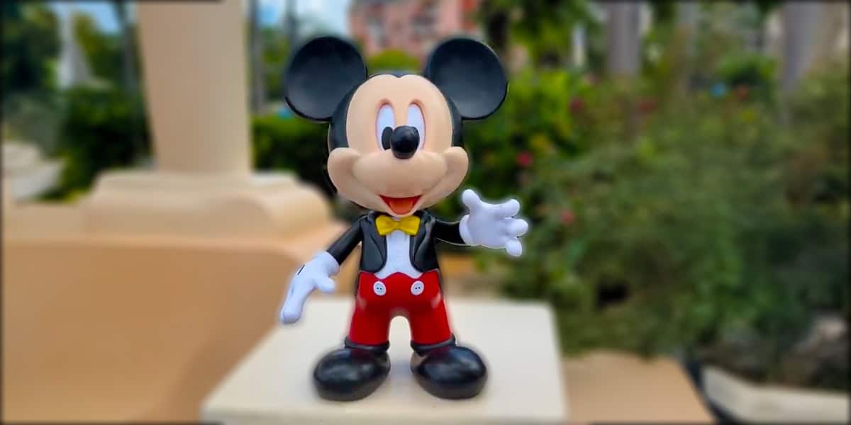 A figurine of Mickey Mouse stands outdoors, waving in his classic red shorts, yellow bow tie, and black shoes. The background is blurred with greenery and a building, evoking the charm of Hollywood Studios.