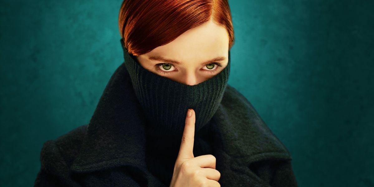 A person with short red hair pulls a dark sweater up over their nose, leaving only their eyes visible, and holds a finger to their lips in a gesture for silence, as if keeping news of the Disney Company sued secret, against a teal background.
