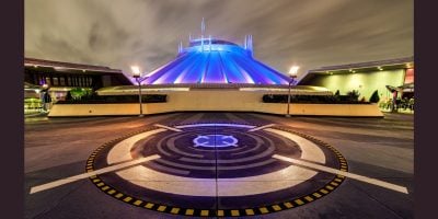 A futuristic, dome-shaped building glows with blue and purple lights at night, set against a cloudy sky. Circular patterns on the pavement hint at a Disneyland Marvel Makeover, while lamps cast a magical glow on the scene.