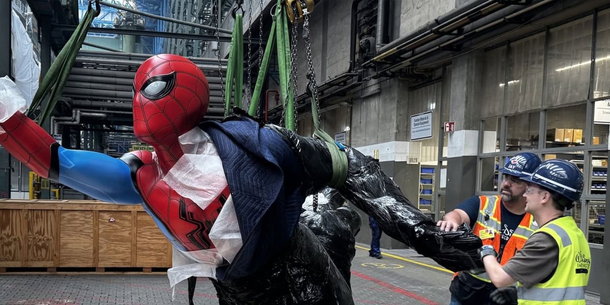 Two workers in safety gear guide a large Marvel Spider-Man figure suspended by green straps in an industrial warehouse, with pipes and equipment visible in the background.