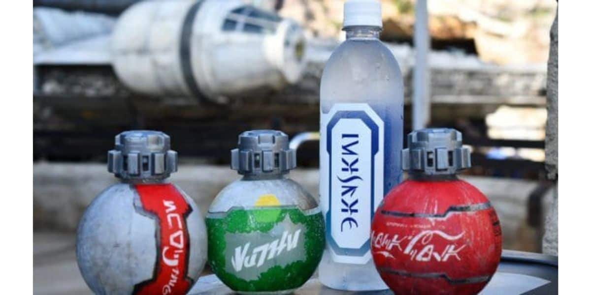 Three round Star Wars Disney Redesign-themed soda bottles and a water bottle with alien script are displayed on a table, with a blurred Millennium Falcon spaceship in the background.