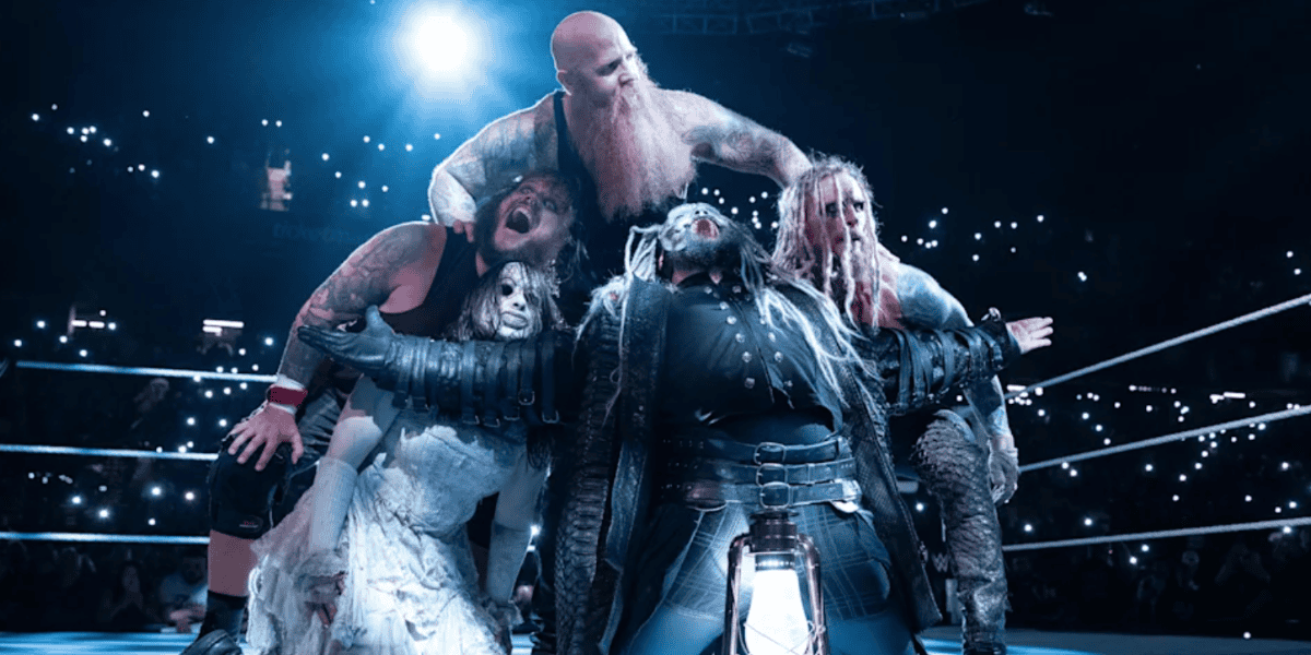 Four wrestlers in dramatic costumes pose in a wrestling ring under spotlight, evoking horror nights vibes, with a lantern at their feet and a crowd holding glowing phones in the dark background.