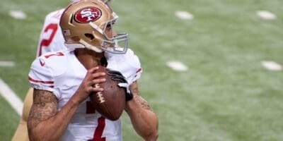 A San Francisco 49ers football player in a white uniform and gold helmet prepares to throw a football during a game, holding the ball with both hands and looking downfield.