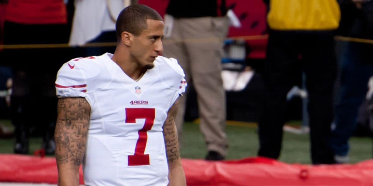 A football player in a white San Francisco 49ers uniform with the number 7 stands on the field, looking to the side. He has short hair and tattoos on his arms. The background shows people and equipment on the sidelines.