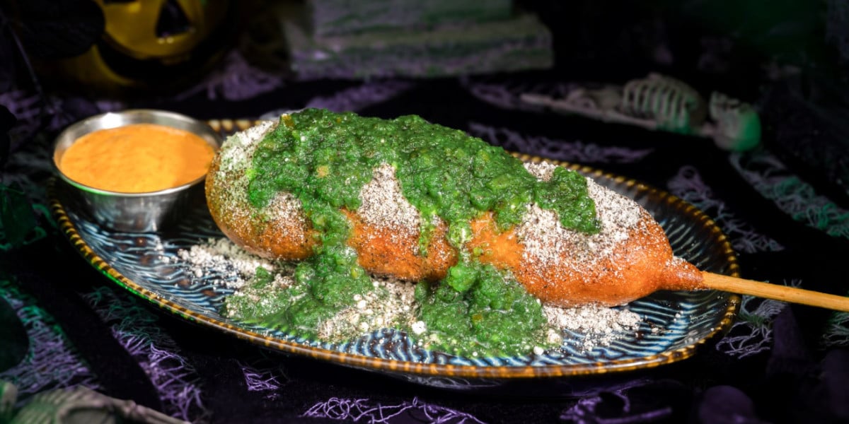 A corn dog on a decorative plate is topped with green sauce and white powder, with a cup of orange dipping sauce on the side. The background features Disney-inspired Halloween decorations.